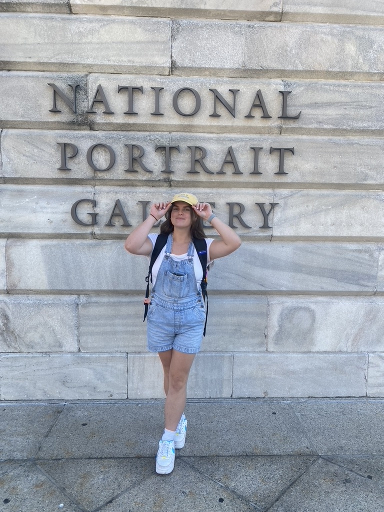 Roxy standing in front of the National Portrait Gallery in Washington DC, wearing blue jean overalls, a white shirt, and a yellow baseball cap that says Art is Activism
