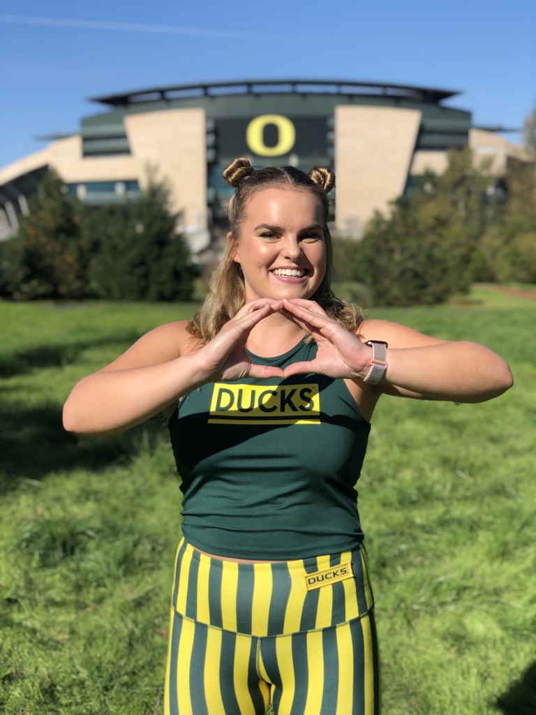 Roxy smiling and making a O shape with her hands in front of Autzen Stadium at the University of Oregon, surrounded by green grass and trees, wearing University of Oregon athletics attire